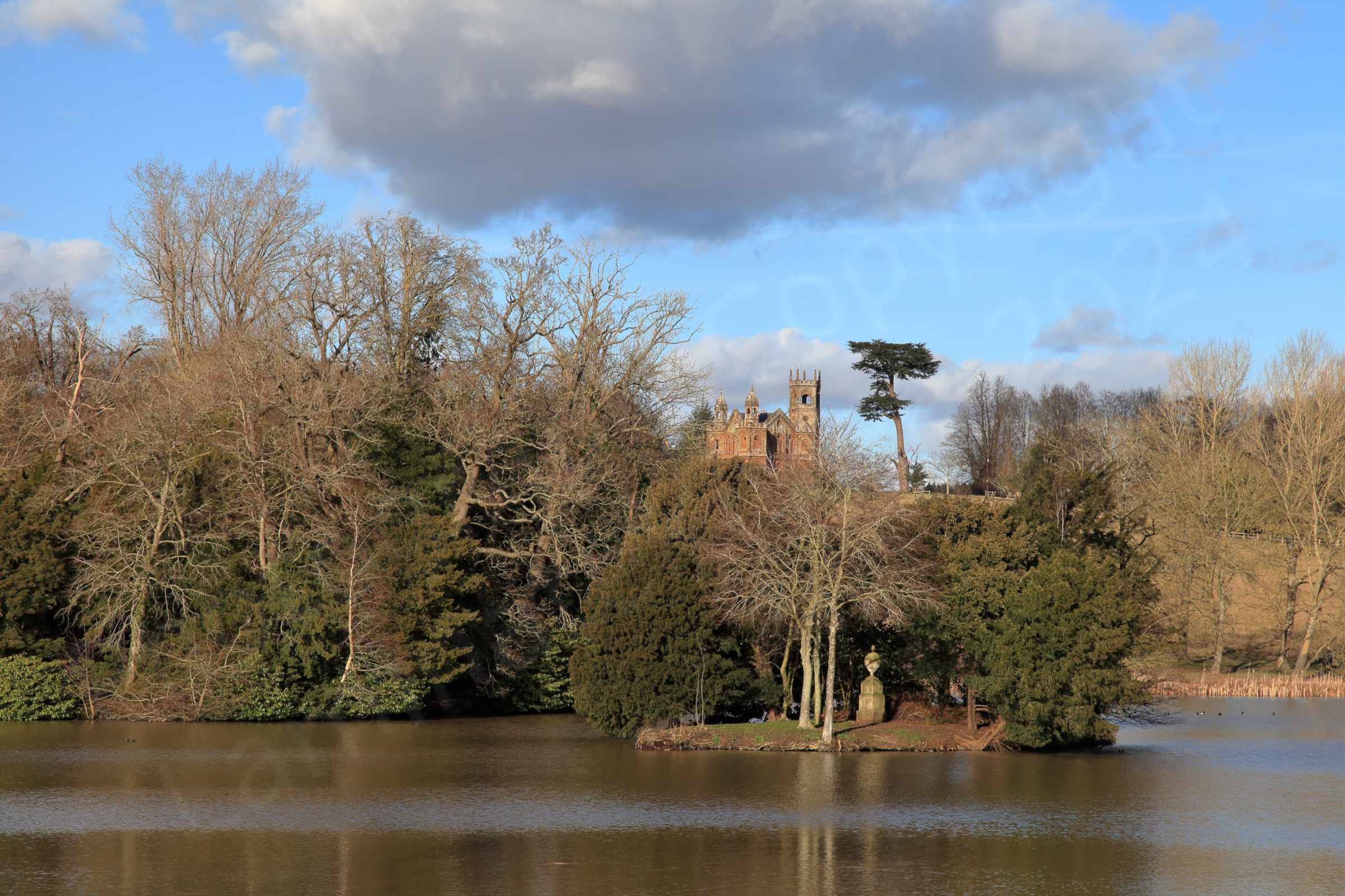 The far side of a narrow stretch of brown water is a slope covered in trees both evergreen and deciduous above which stands the Gothic Temple with its tower and twin belfries.
