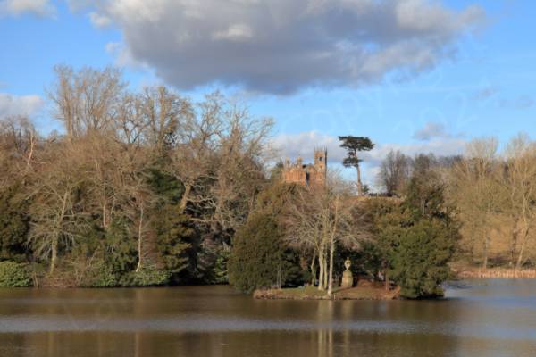 The far side of a narrow stretch of brown water is a slope covered in trees both evergreen and deciduous above which stands the Gothic Temple with its tower and twin belfries.